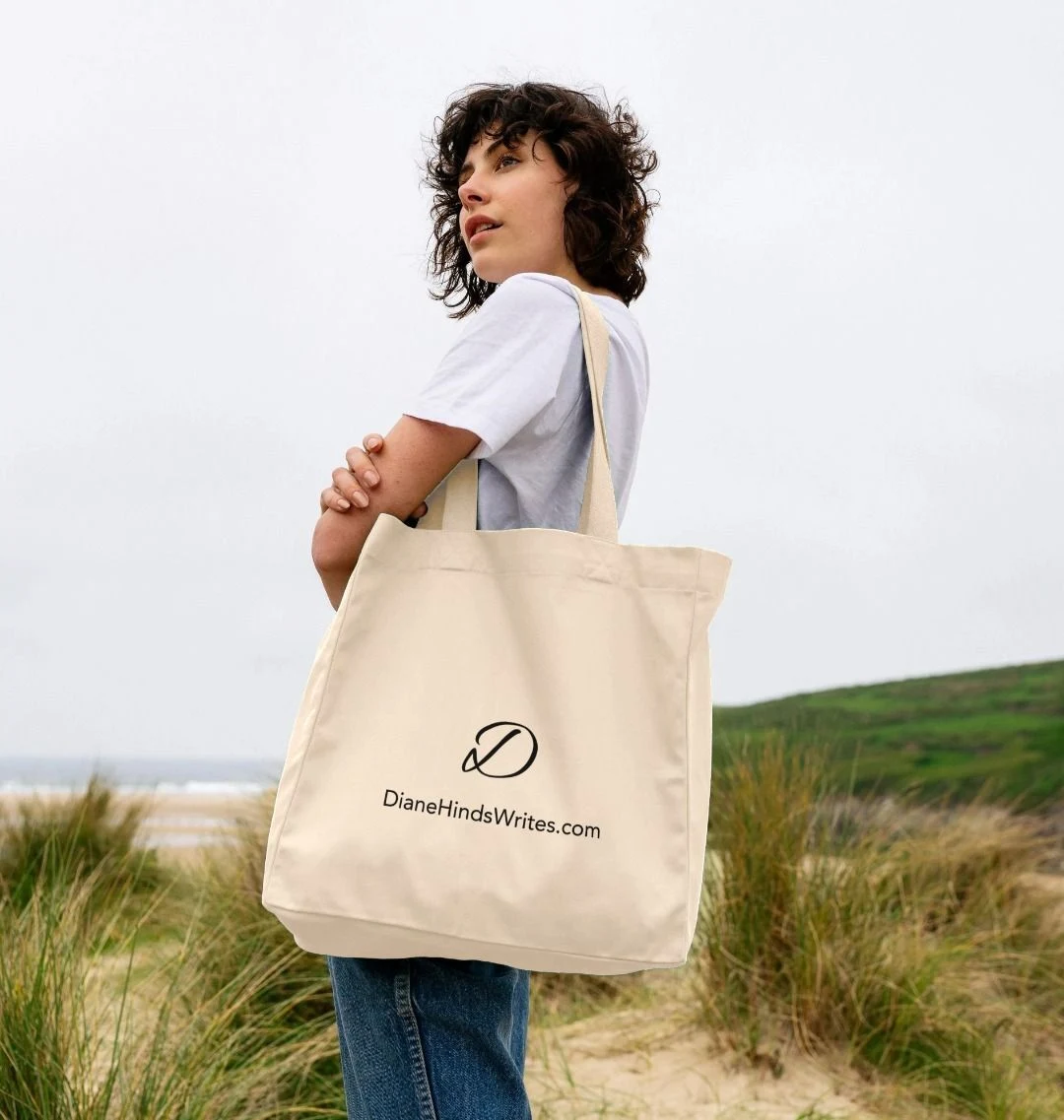 Model holding a natural-coloured organic cotton tote bag featuring the DianeHindsWrites.com logo, standing on a sandy beach with grassy dunes